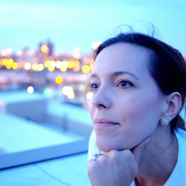 Travel Advisor Sloane Davidson looks into the distance on a rooftop with a sparkling city at sunset behind her 