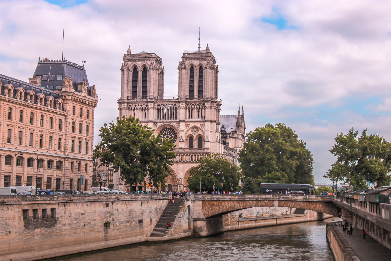 A large tan church - the Notre Dame in Paris - with stained glass windows two pillars and a bridge over a river with green trees