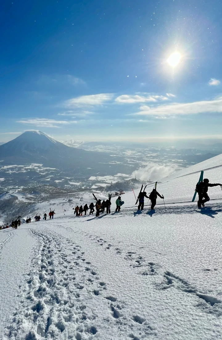 A group of people trekking up a snowy mountain with another large mountain in the far distance under the shining sun. 