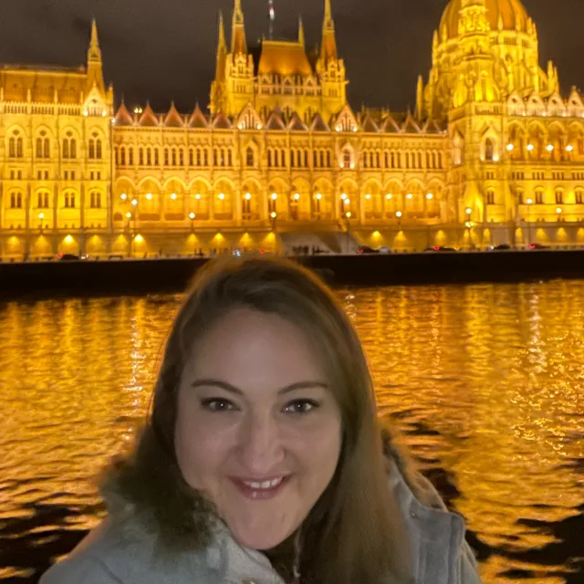 Travel Advisor Meg Lake smiles in front of a castle on the water brightly lit up by golden light