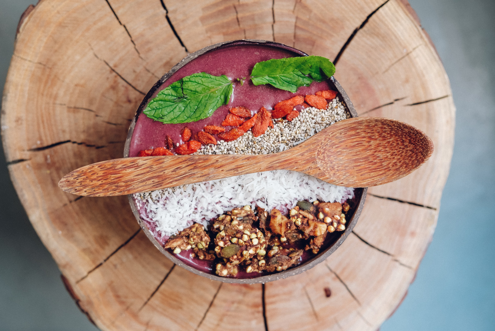 Wood bowl with pink acai, white coconut and granola with a wooden spoon on top on a wood table
