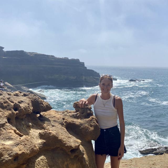 Fora travel agent Katie Bernal-Silva wearing white tank top and black shorts stands next to rock with ocean in background