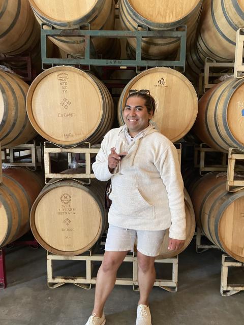 Fora travel agent Gregory Rodriquez wearing white shirt holding drink in front wine barrels