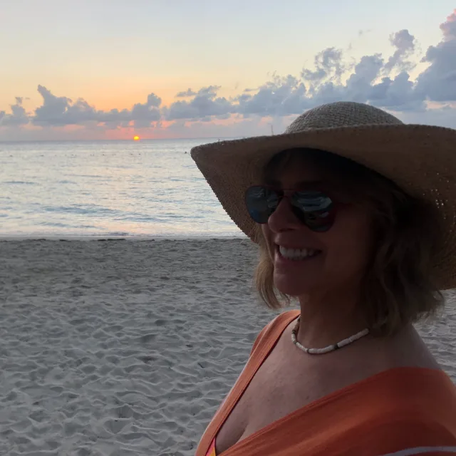 Travel Advisor Karen Brucki Ziliak in an orange top and hat, enjoying sunset on a beach. 