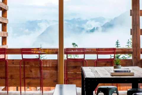 A black table and red stools against a wooden ledge with mountains and clouds in the background. 