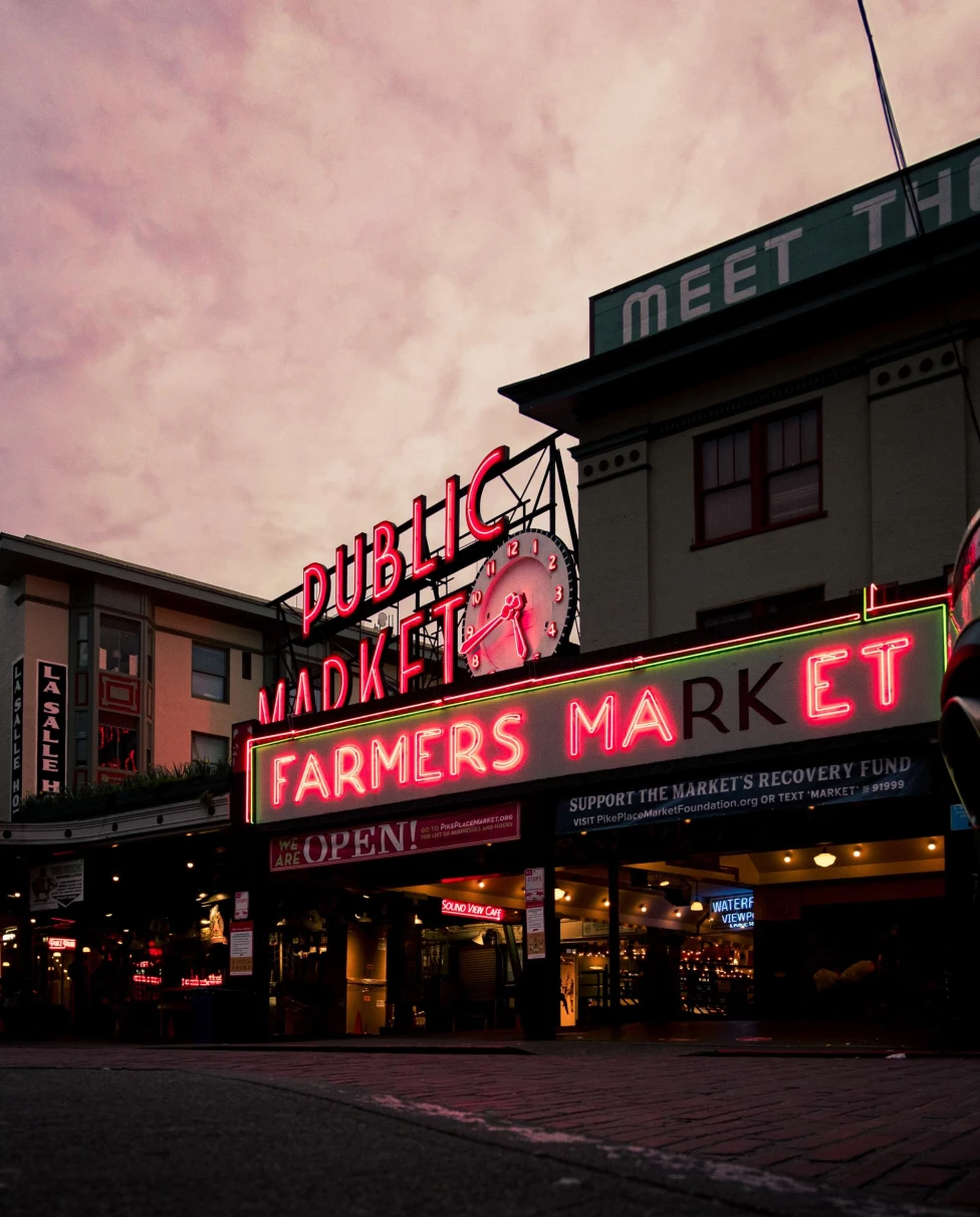 neon sign reads, "public market" and "farmers market" under purple sunset