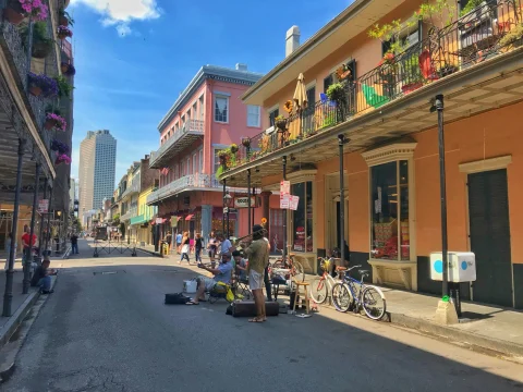 A view of a vibrant street full of people in New Orleans. There are also bicycles and orange and pink buildings with metal balconies, decorations and plants in the surrounding areas. 