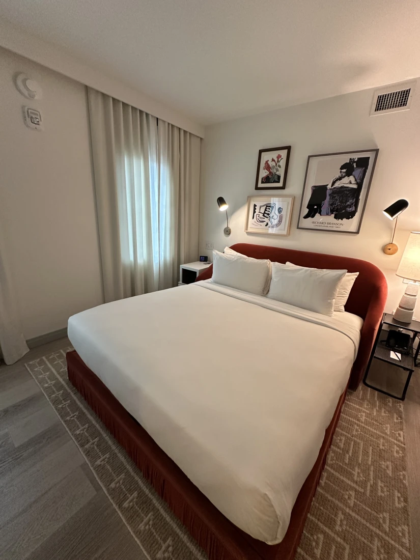 A bed in the hotel room with white bedding and a red base and headboard, with white walls and artwork above the bed.