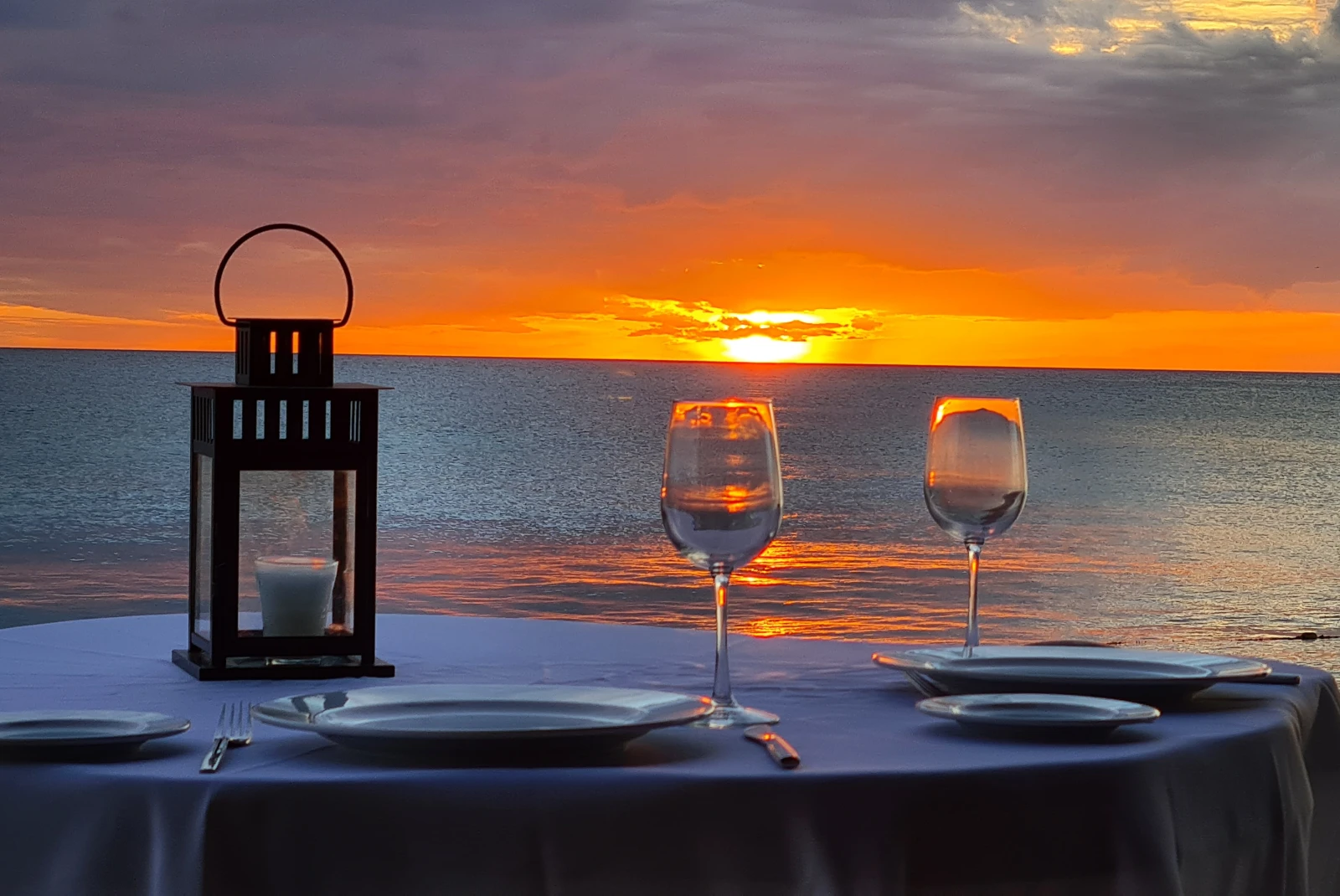 Two wine glasses sit on a table with a blue table cloth overlooking the ocean at sunset