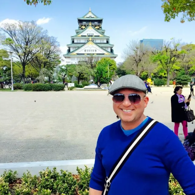 Travel Advisor Travis Tanner wears a blue sweater, cross body bag, sunglasses, and a beret hat in front of a Japanese temple. 