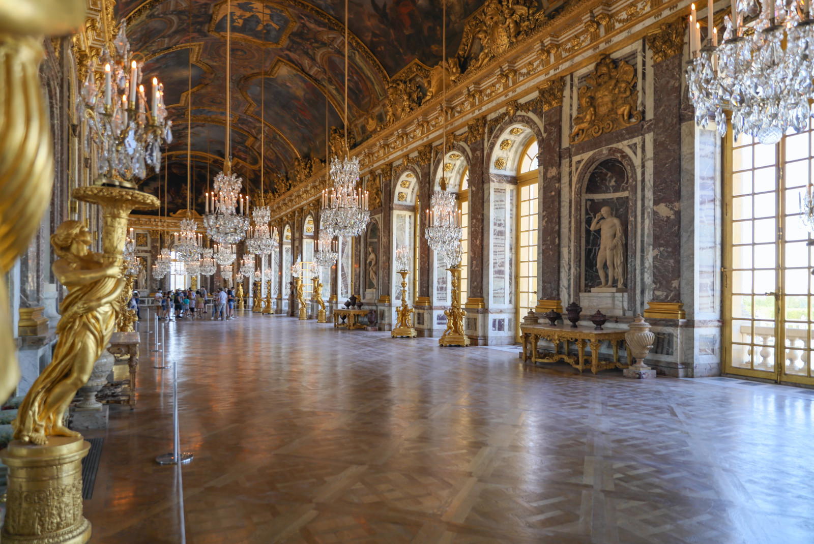 A large open hallway with hanging glass chandeliers and gold and white statues with paintings covering the ceiling