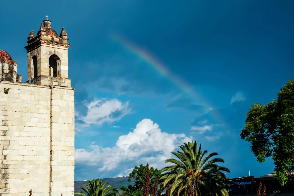 a blue sky with a rainbow and palm trees in front of an ancient church