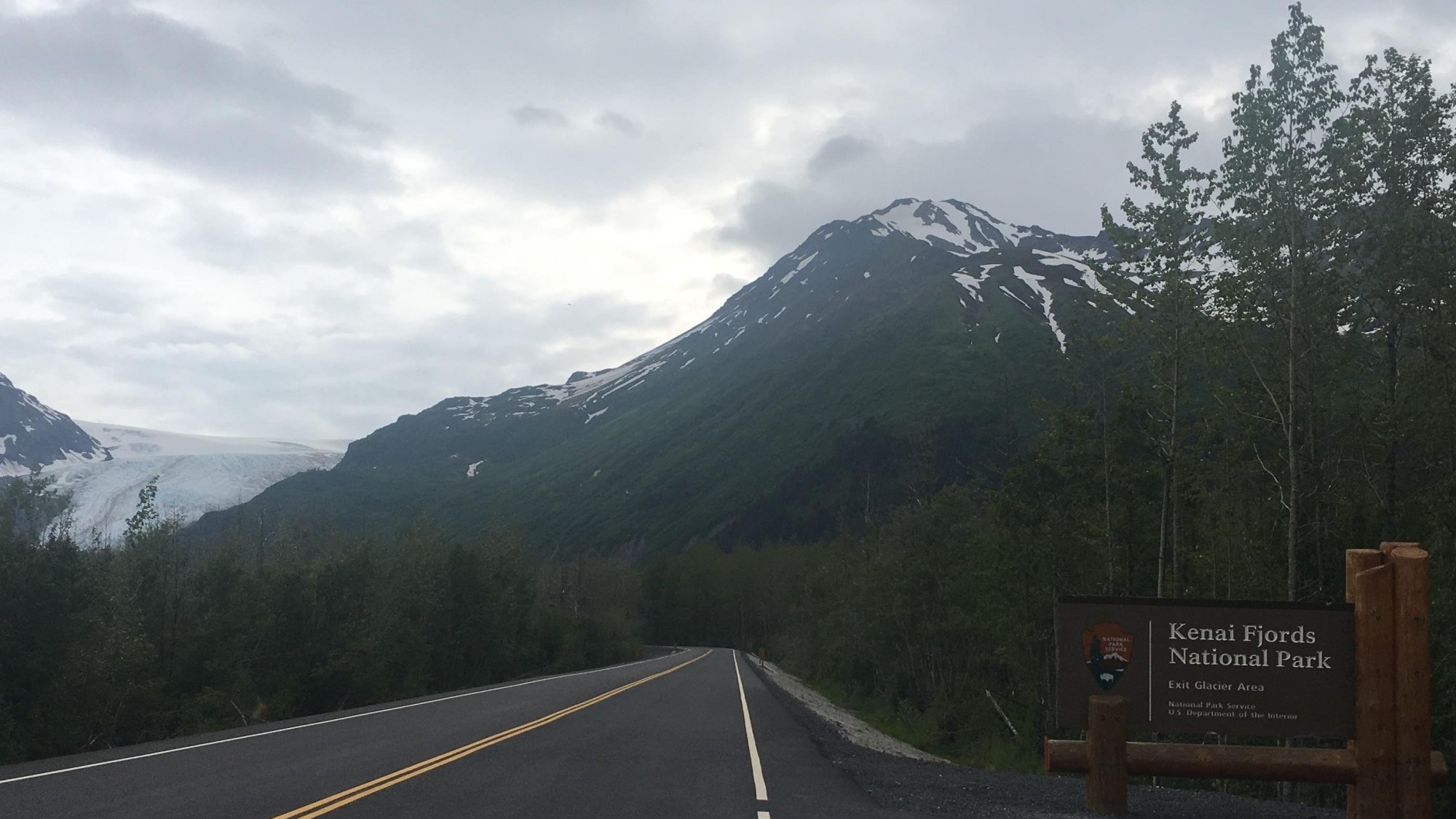 Road with snow covered mountains at the back and board saying Kenai Fjords National Park. 