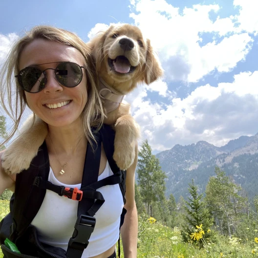 Travel Advisor Charlie Gaffney smiles at the top of a wild flower mountain with a black backpack and a dog smiling over her shoulder