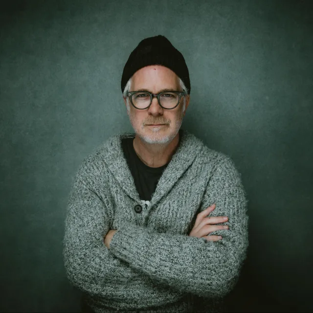 Picture of Michael in grey sweater with his arms crossed standing in front of a teal wall.