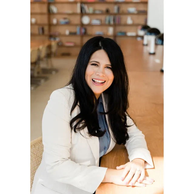 Picture of Alejandra in a white suit sitting at a table with a bookshelf faded in the background