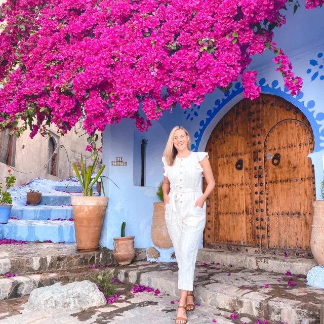 Travel Advisor Michelle Cassidy stands in front of a vibrant pink flowering plant on a pastel blue building with a arched wood door wearing white 