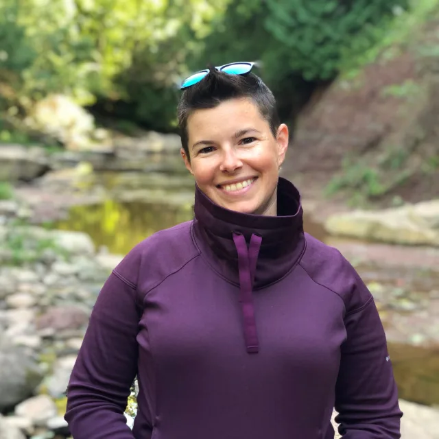 Travel Advisor Sherry Buck in purple jacket and sunglasses on head in front of a river.