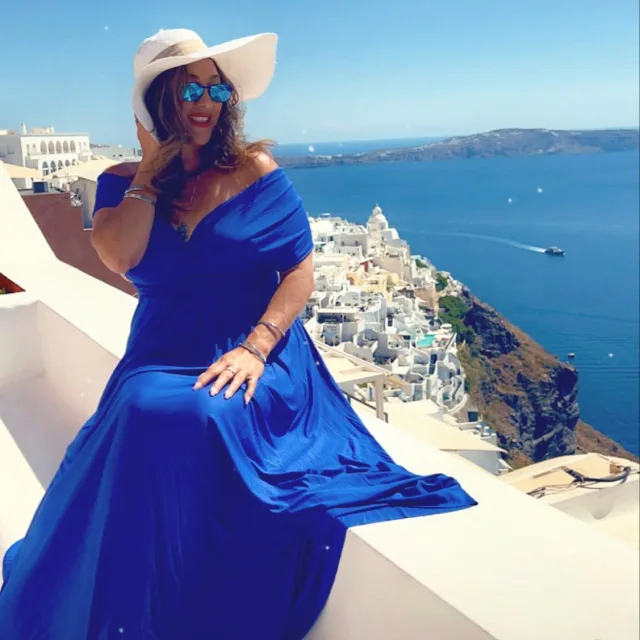 Travel advisor Christie Ann Mitsumura in Greece sitting on the wall wearing Blue dress and off-white hat.