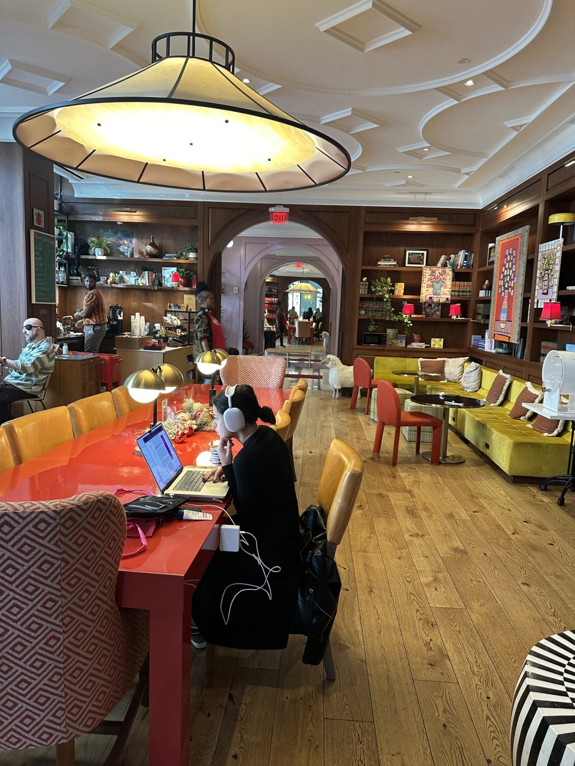 Lounge area at the Funny Library Coffee Shop with one woman working on a laptop and others sitting. There is a long red table and a yellow long sofa in view, as well as artwork and accessories on the shelves and walls.
