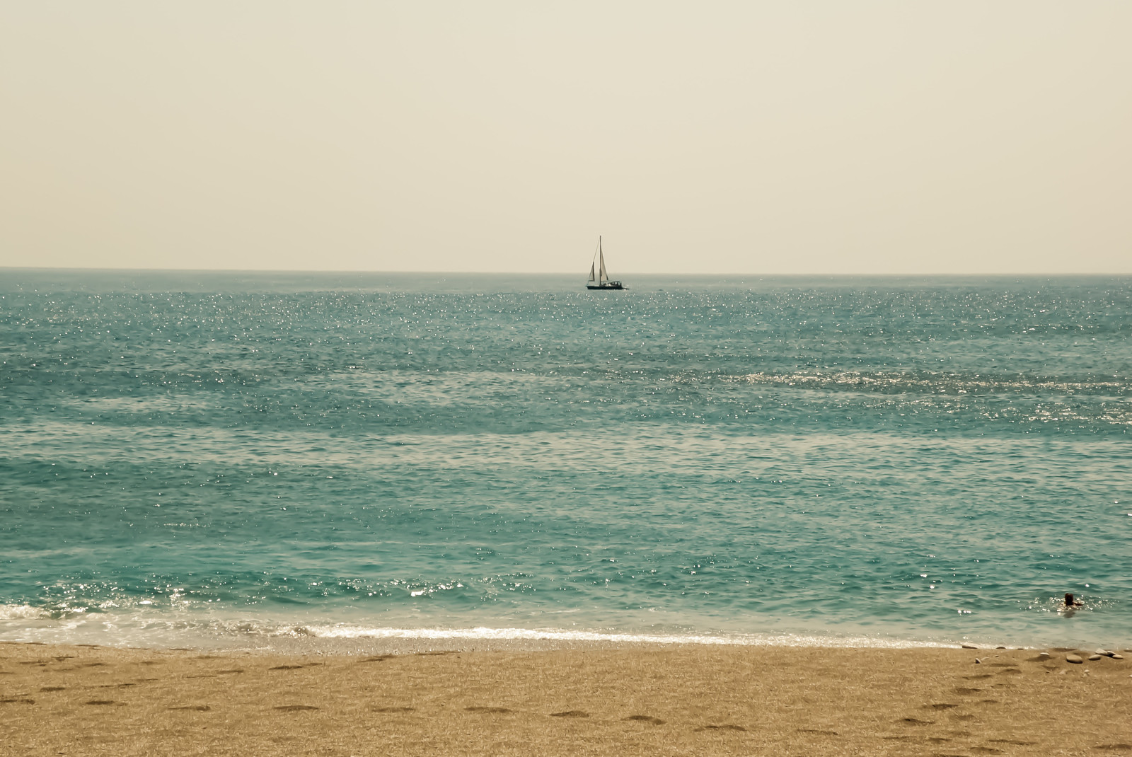 blue water with white waves and a sailboat tan sand with footprints