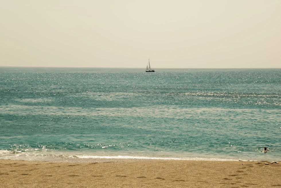 blue water with white waves and a sailboat tan sand with footprints
