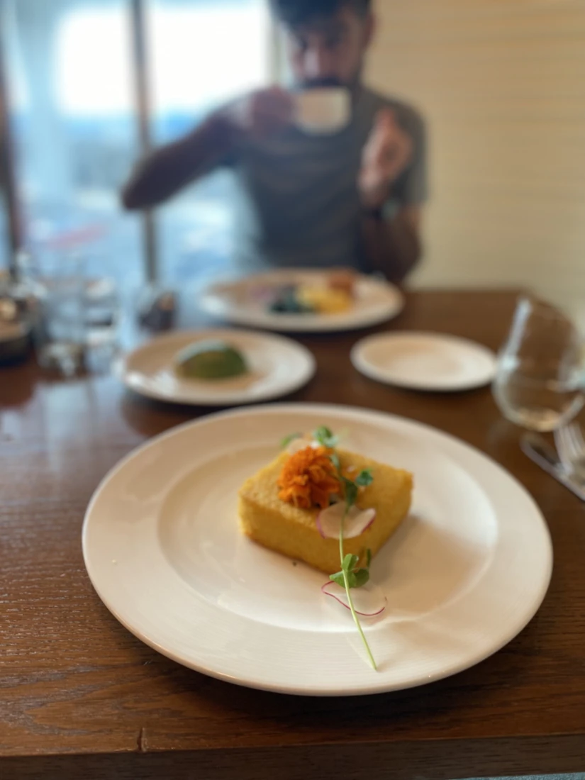 A view of a yellow pastry on top of a white plate. There is a person sipping coffee out of focus in the background. 