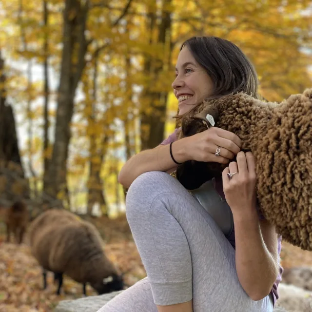 Travel Advisor Jamaica Kelley in a gray outfit hugging a brown sheep with yellow leaved trees.