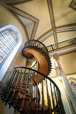 The image features an ornate spiral staircase with intricate railings beside stained glass windows, within a classically styled interior.