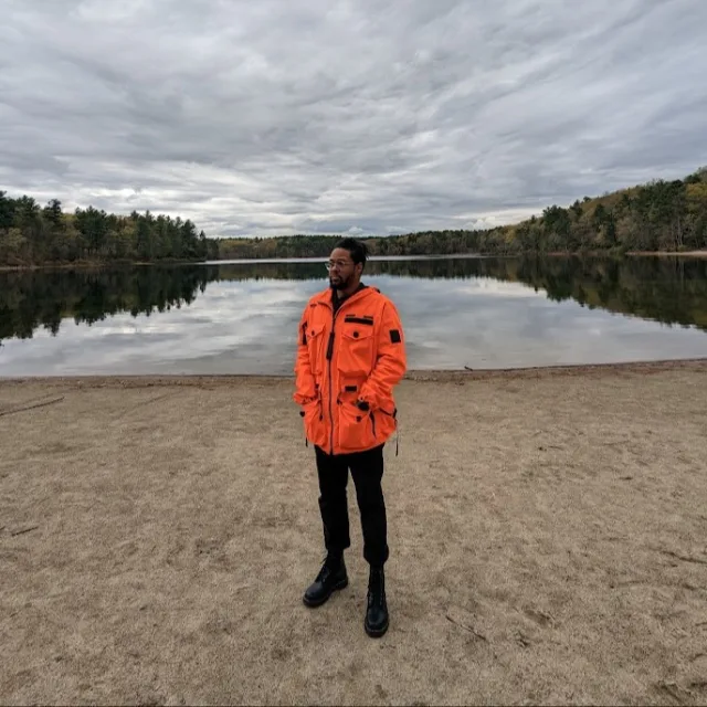 Norris Howard in an orange jacket posing on a beach with trees and water in the background on a cloudy day