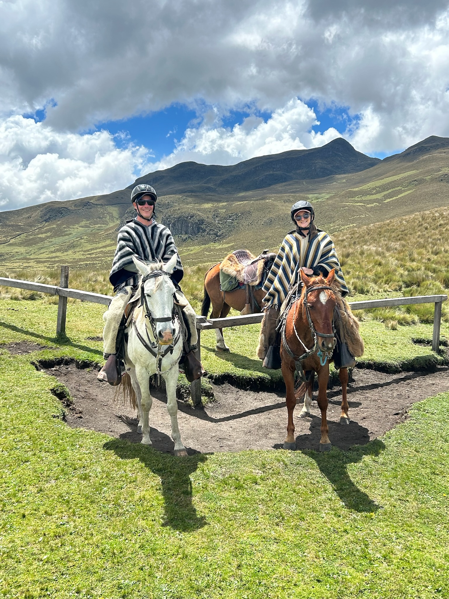 Stephen and his partner each riding a horse while wearing a black and white striped poncho outside on a green meadow with a mountain range in the background