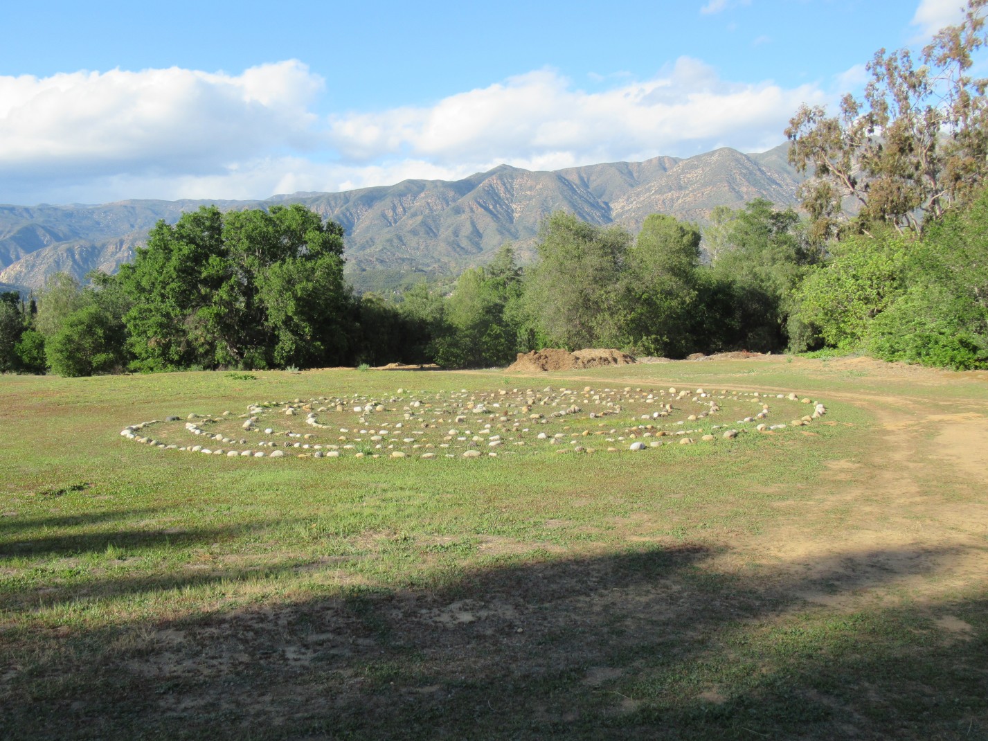 Rocks laid out in a circle for meditation practice on green grass surrounded by trees with mountains in the background and a blue sky