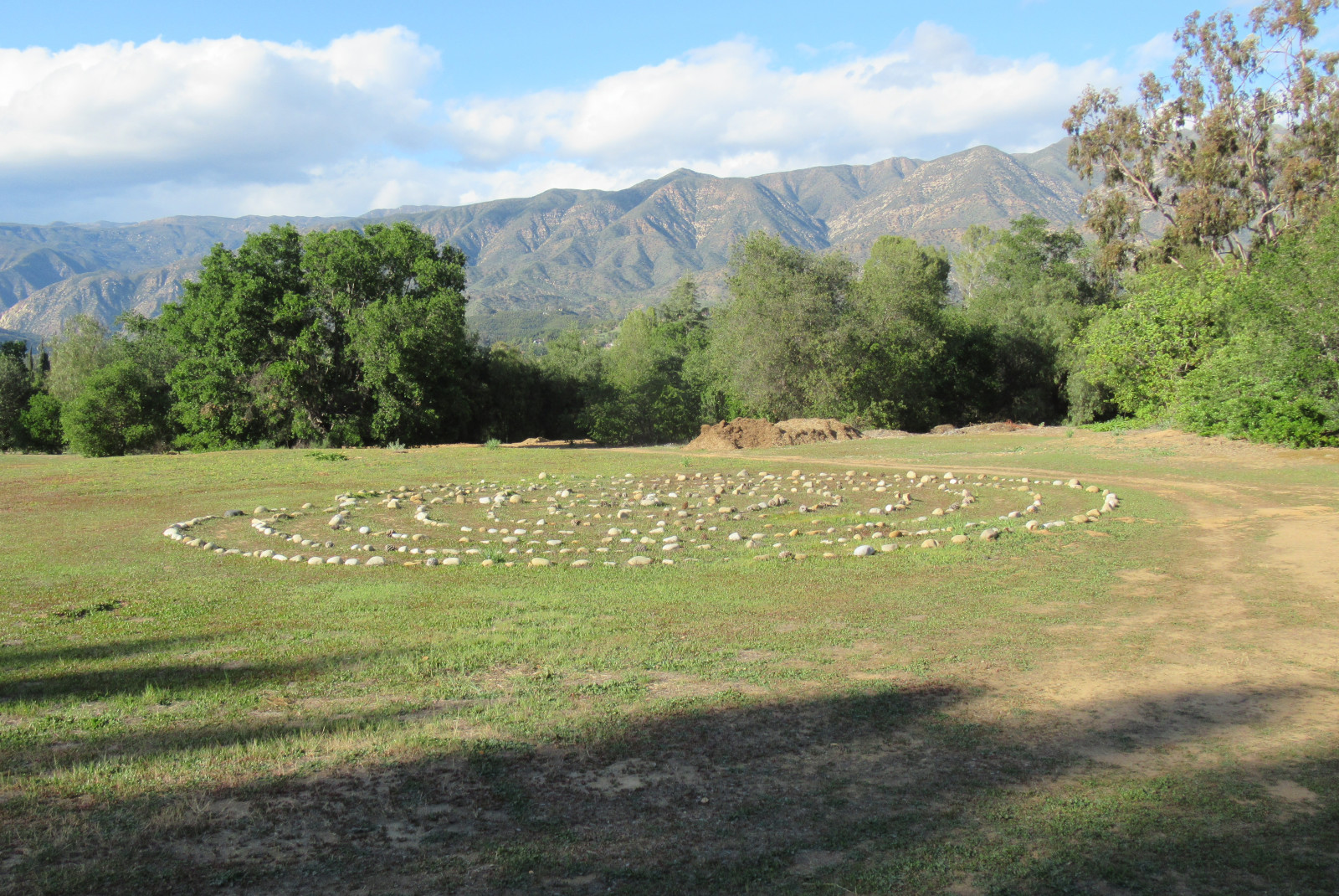 Rocks laid out in a circle for meditation practice on green grass surrounded by trees with mountains in the background and a blue sky