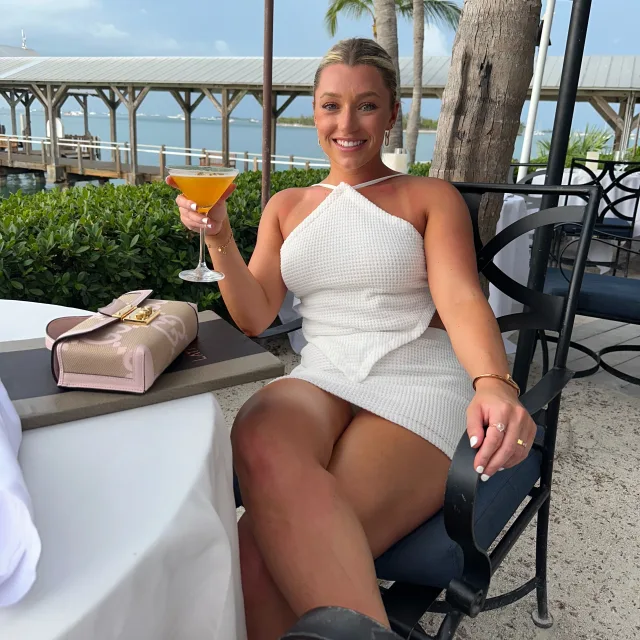 Zoe wearing a white dress and holding up an orange cocktail while seated outside at a dinner table with trees, a boardwalk and water in the background