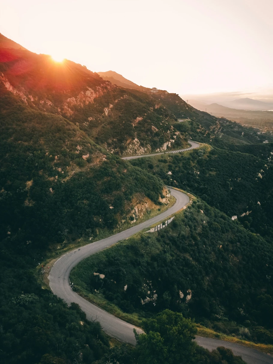 An aerial view of a winding road through green mountains during the sunrise.