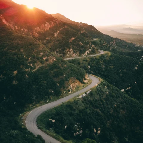 An aerial view of a winding road through green mountains during the sunrise.