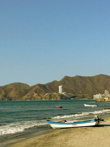 A view of a small boat on a sandy beach shore with the blue water, two boats, city shoreline and brown mountains in the background. 