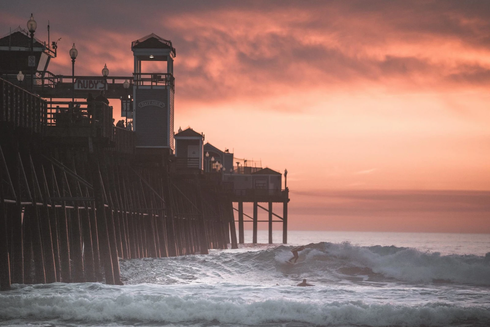 Waves crashing on legs of the pier in Oceanside during cloudy orange sunset.