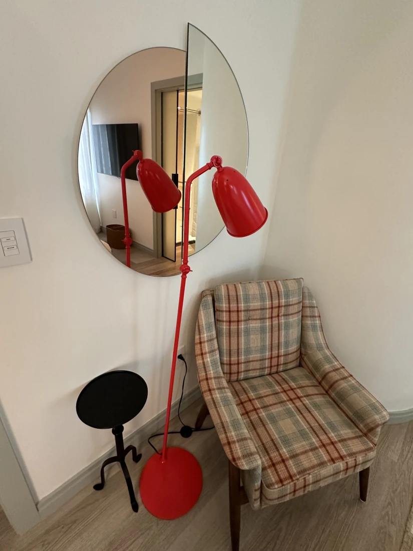 A plaid arm chair with a bright red floor lamp, small black table and large round mirror in the hotel room