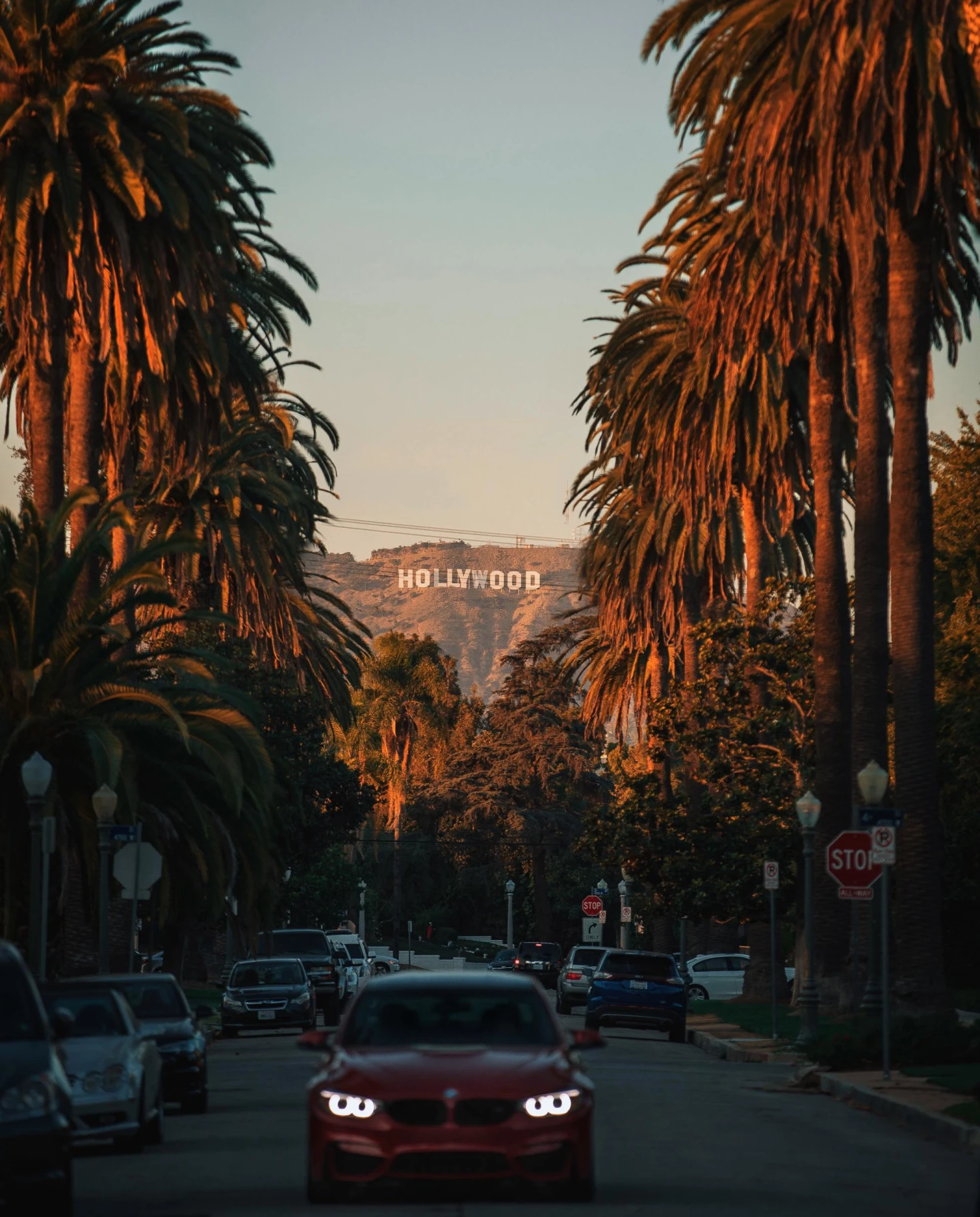 a red car drives down a road lined with palm trees at sunset with the Hollywood sign in the distance