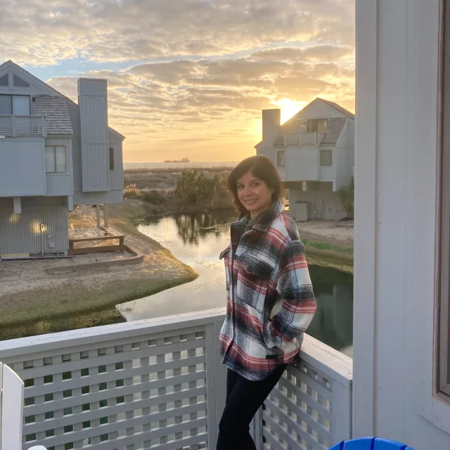 Travel Advisor Gail Graham in a plaid sweater in front of houses and water at sunset.
