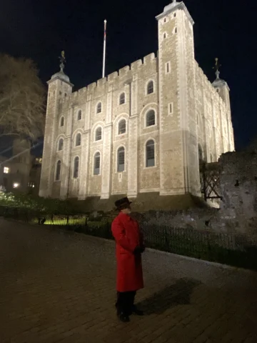 A man in red coat and hat standing in front of a well lit building at night.