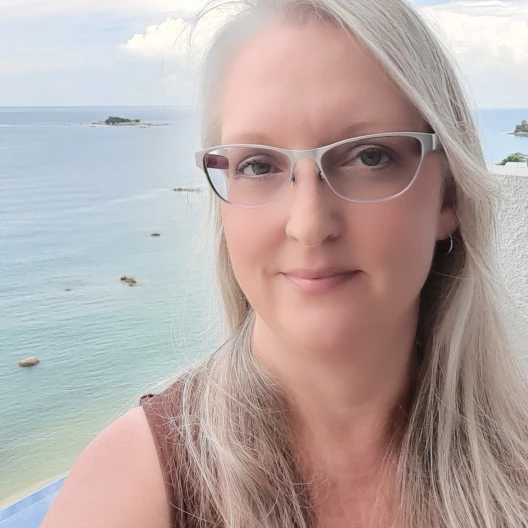 Fora Shella Zelenz Travel Agent wears grey rimmed glasses and brown shirt with ocean in the background