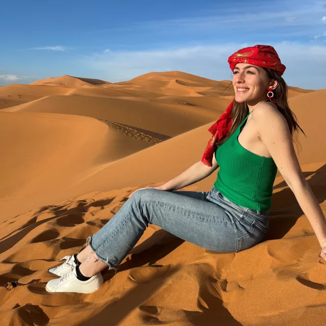 Stephanie posing in the desert sand dunes while wearing jeans, a green top and a red scarf on her head.