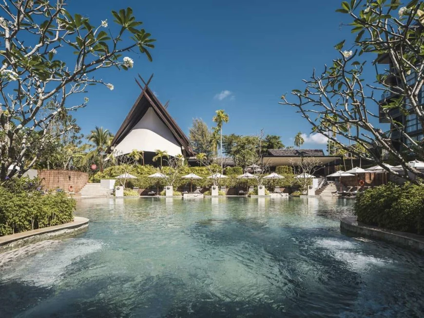 a thatched-roof building behind a sprawling swimming pool