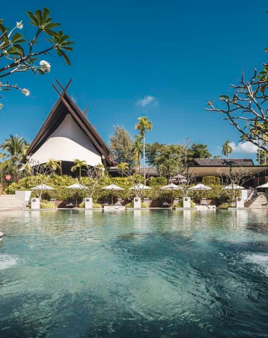 a thatched-roof building behind a sprawling swimming pool