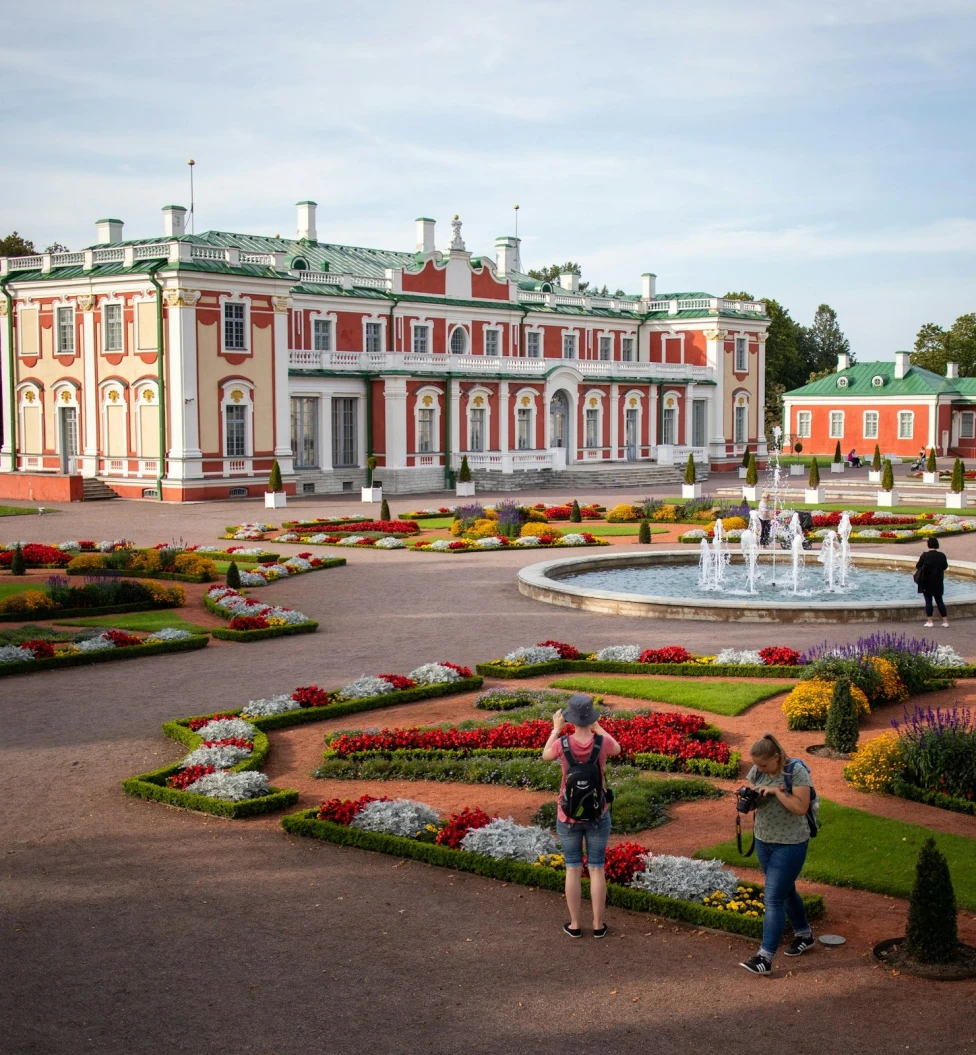 Kadriorg Park is the most outstanding palatial and urban park in Estonia. Picture features manicured gardens and a circular fountain in front of a large red building with white trim and green roofs.