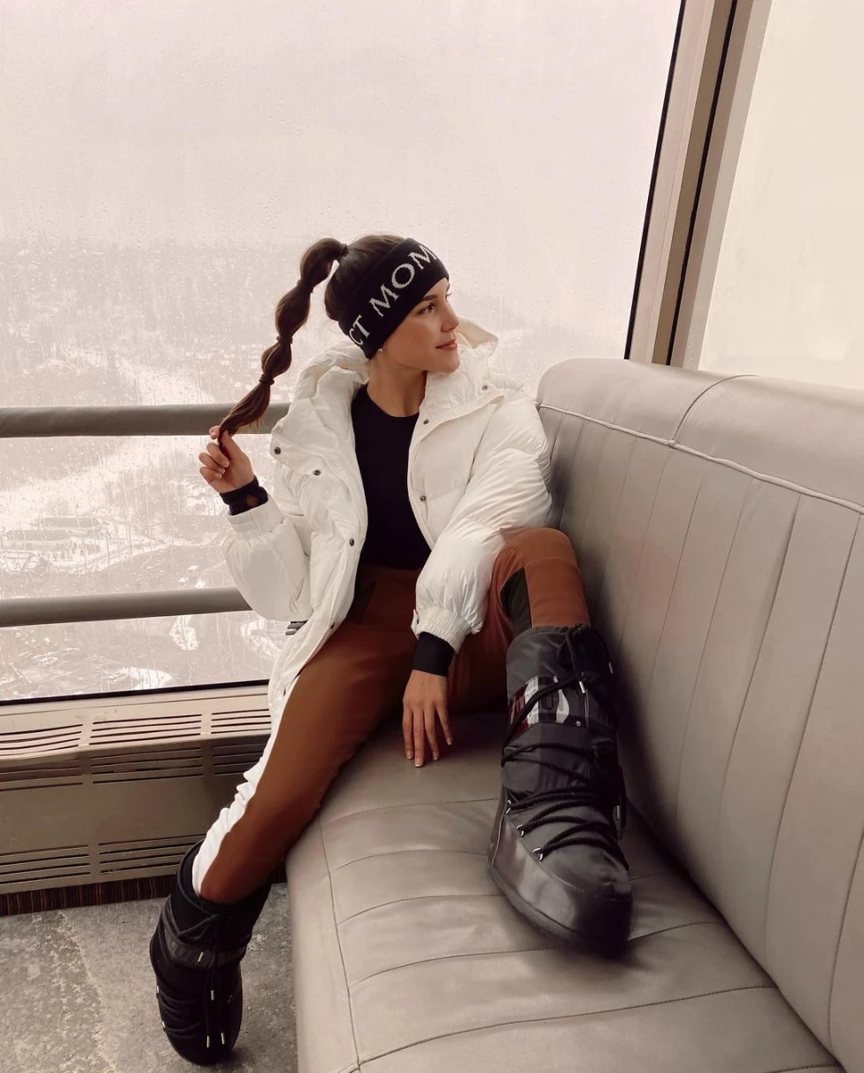 A lady in posing in white jacking and long leather boots with a an aerial view of snow c;lad valley at the background. 