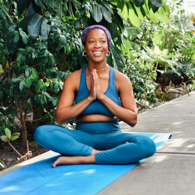 Woman wearing blue pants sitting on yoga mat with green foliage in background