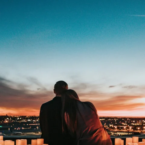 A silhouette of a couple posing while watching the sunset with candles lit around them and a city view in the distance. 